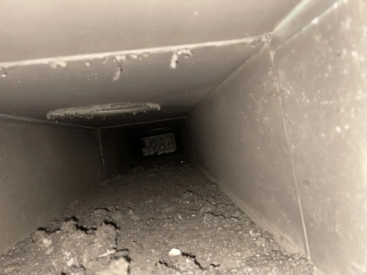 BEFORE air duct cleaning: steel return trunk with thick floor-level debris buildup visible from the opening