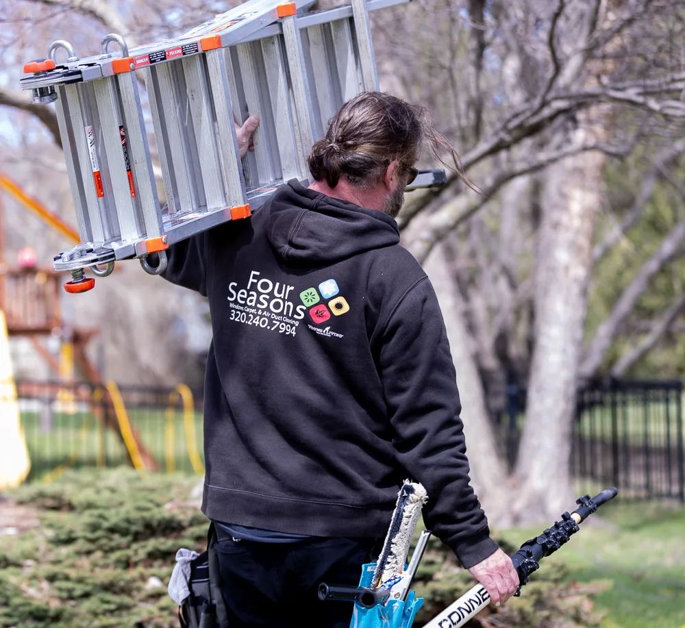 Matt Bertsch carrying a ladder over his shoulder with window cleaning equipment, Four Seasons logo visible on hoodie back