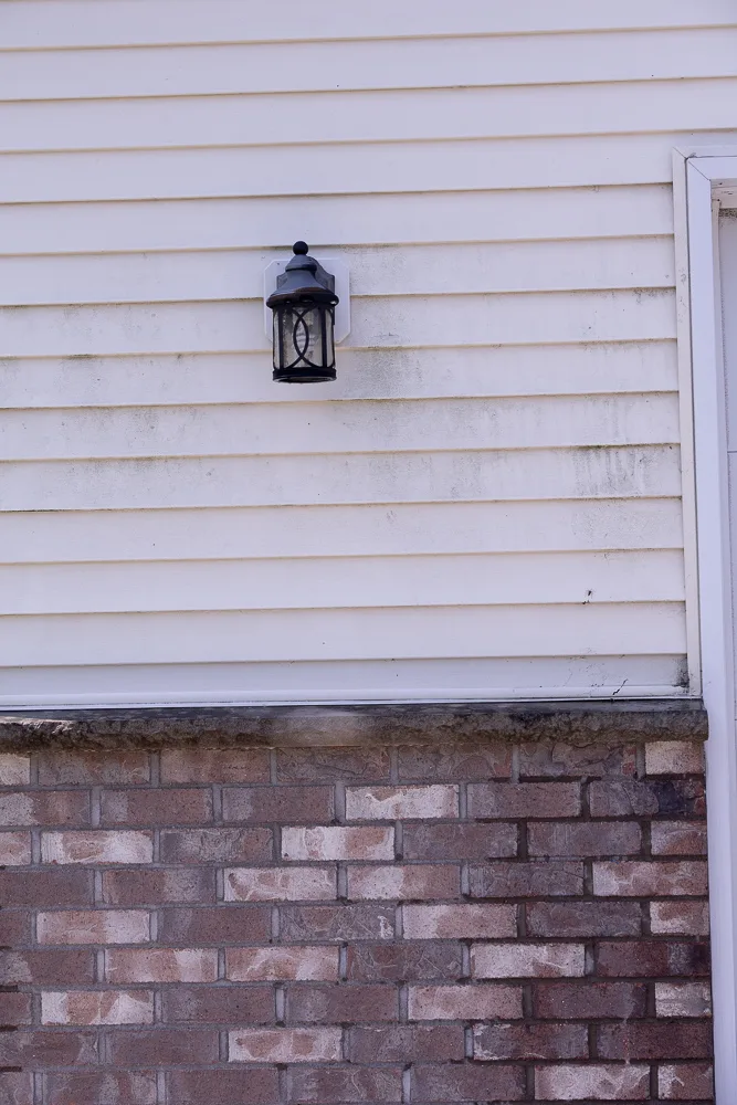BEFORE soft washing: green algae and grime streaking vinyl siding above a brick foundation