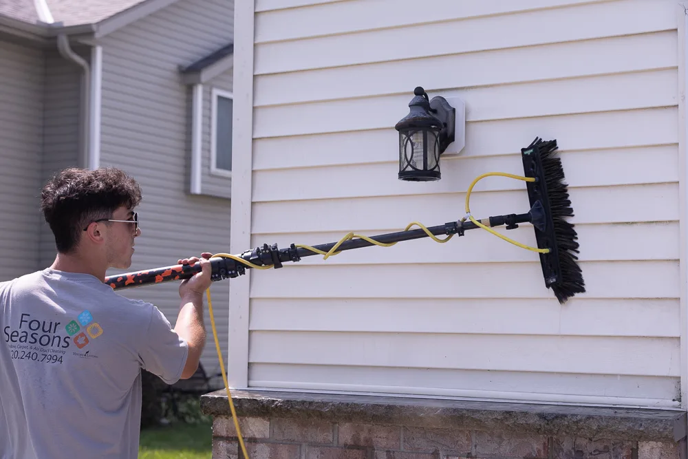 Four Seasons technician in branded company shirt soft washing siding near a light fixture with a soft-bristle brush