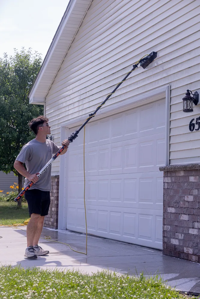 Four Seasons technician reaching a water-fed pole and brush up to soft wash siding above a garage door