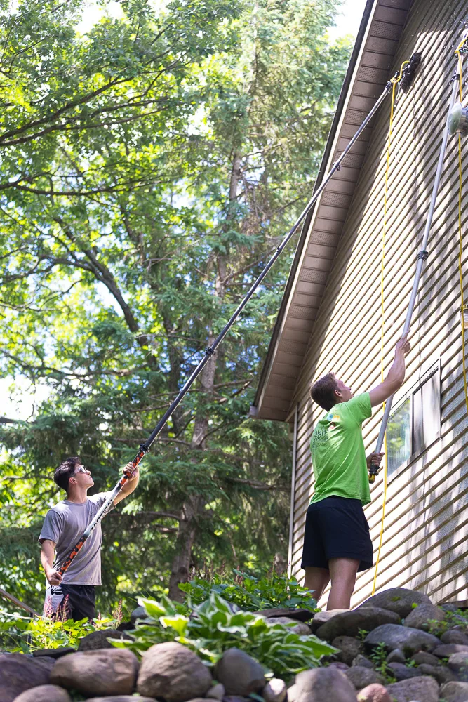 Two Four Seasons technicians soft washing siding along a home's roofline with water-fed poles